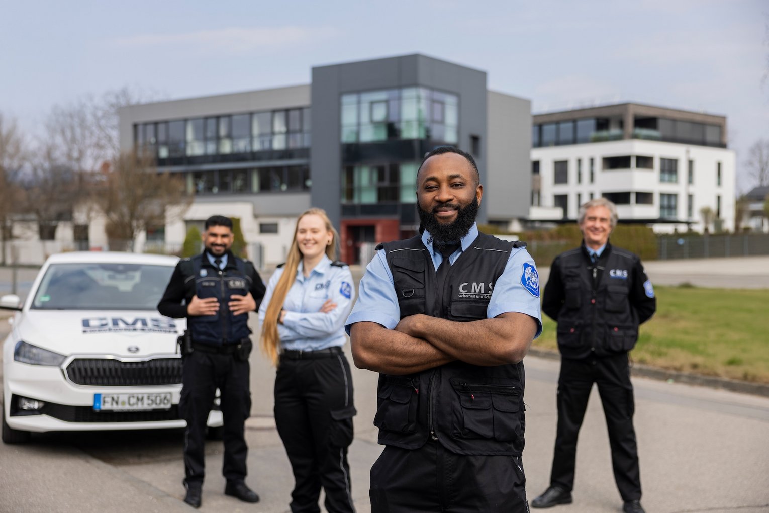 Sicherheitsdienst-Team von CMS steht vor modernem Bürogebäude, Mitarbeiter in Uniform mit Einsatzfahrzeug im Hintergrund