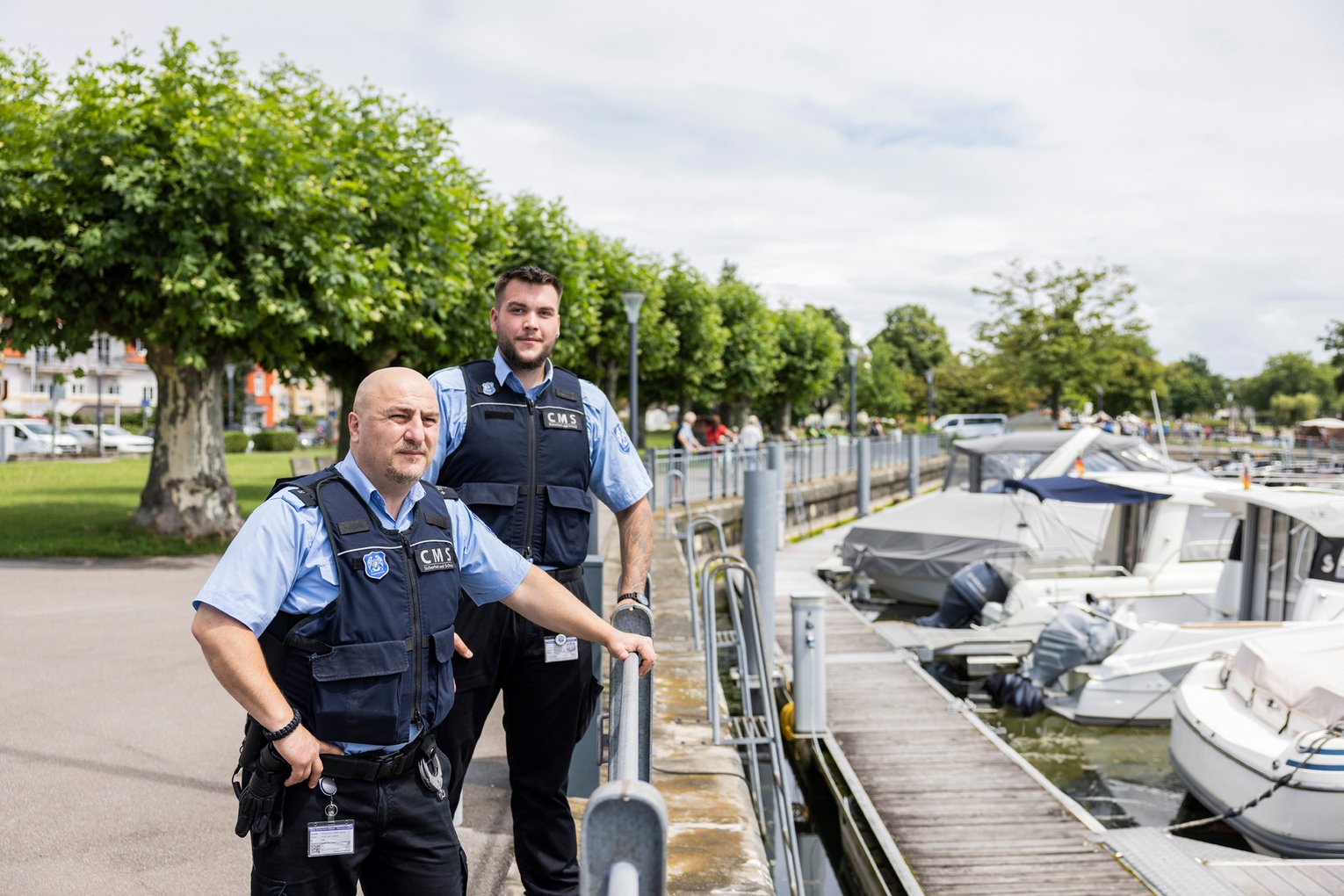 Zwei Sicherheitsdienst-Mitarbeiter in Uniform patrouillieren am Ufer eines Hafens mit Booten, im Hintergrund Bäume und ein Gehweg.